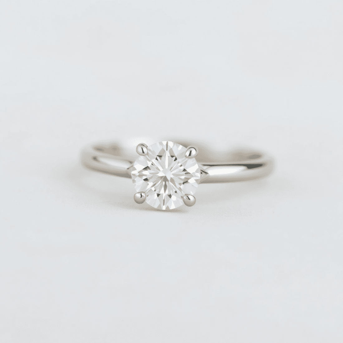 Close-up of a sparkling solitaire diamond engagement ring with a simple band on a soft, neutral background, highlighting the gemstone's brilliance and elegance.