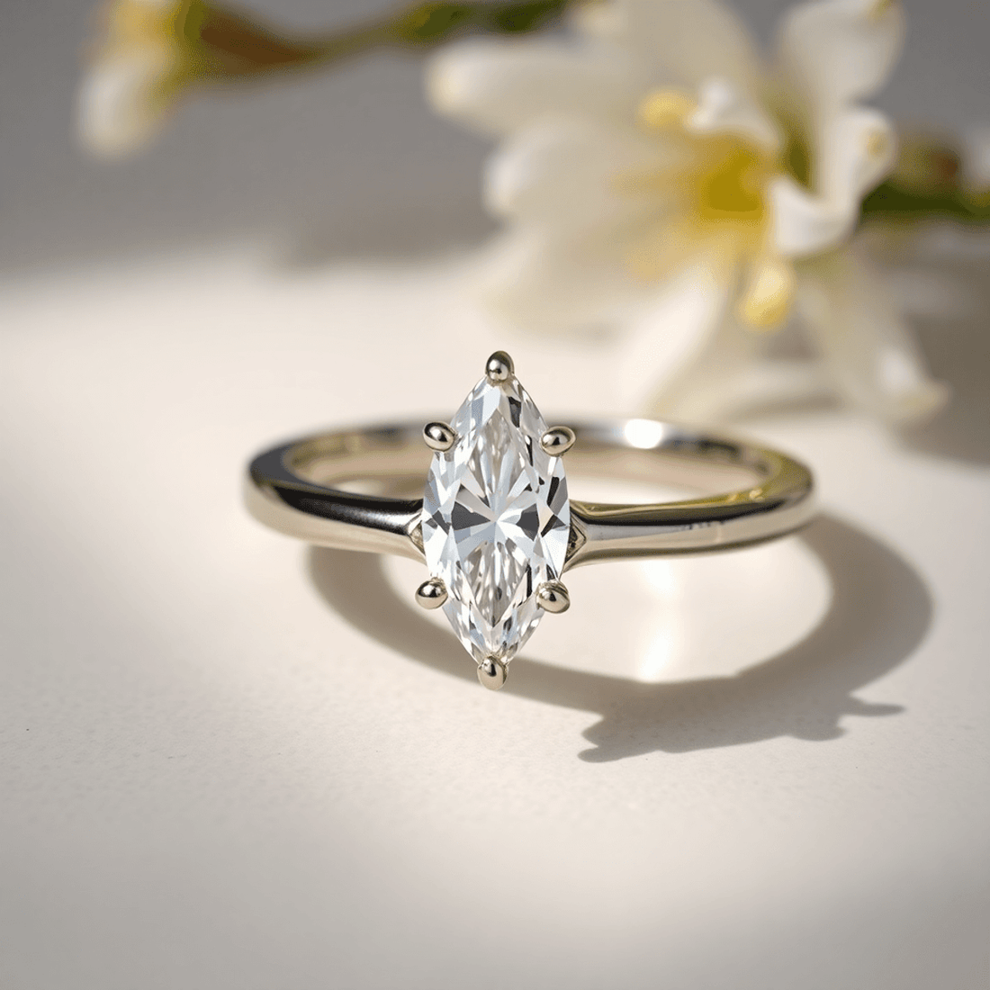 Close-up of a sparkling marquise cut diamond ring on an elegant band, highlighted by soft natural light with subtle floral accents on a neutral background.