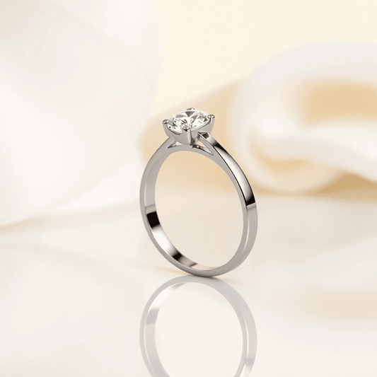 Close-up of a sleek engagement ring with a polished band and sparkling gemstone on a reflective surface with soft neutral background.
