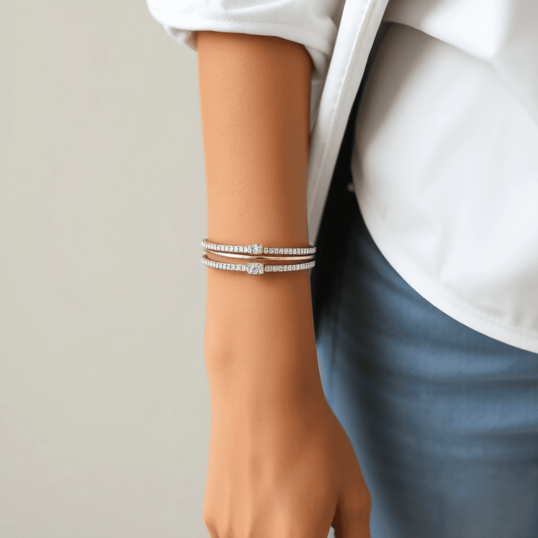 Close-up of a wrist wearing delicate diamond bangles paired with a white shirt and denim jeans against a soft, neutral background.