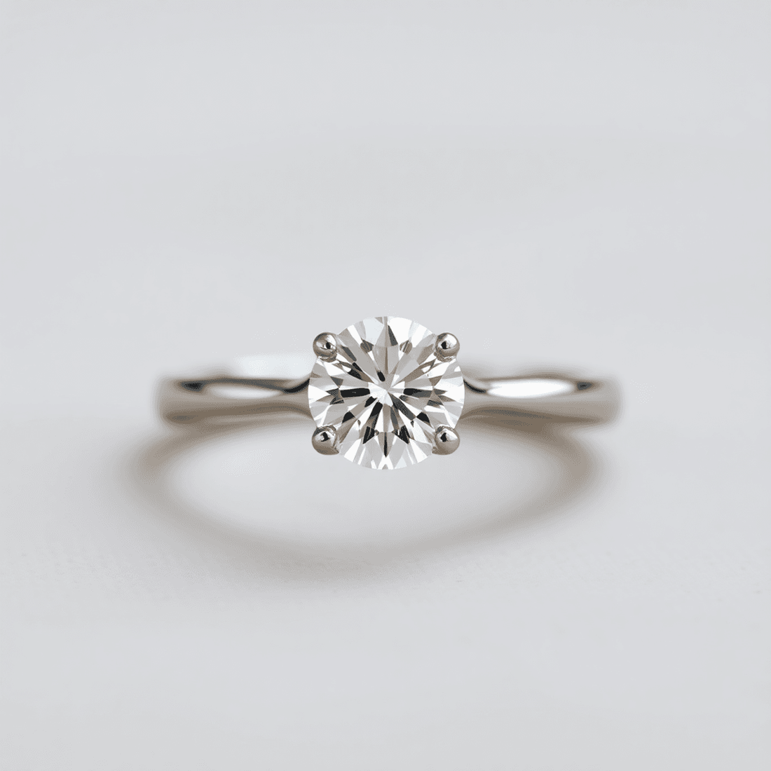Close-up of a sparkling round brilliant diamond solitaire ring on a sleek platinum or gold band against a soft, neutral background with gentle lighting.