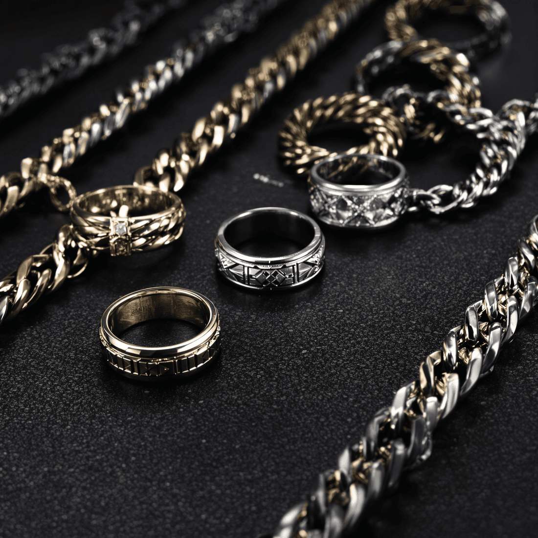 Close-up of men's silver and gold rings and chains on a dark textured surface, showcasing a modern and sophisticated style.