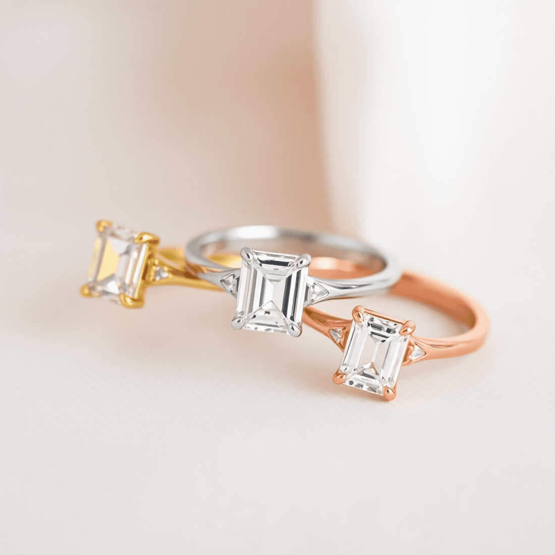 Close-up of three emerald cut diamond rings in yellow gold, white gold, and rose gold on a soft neutral background with gentle lighting.
