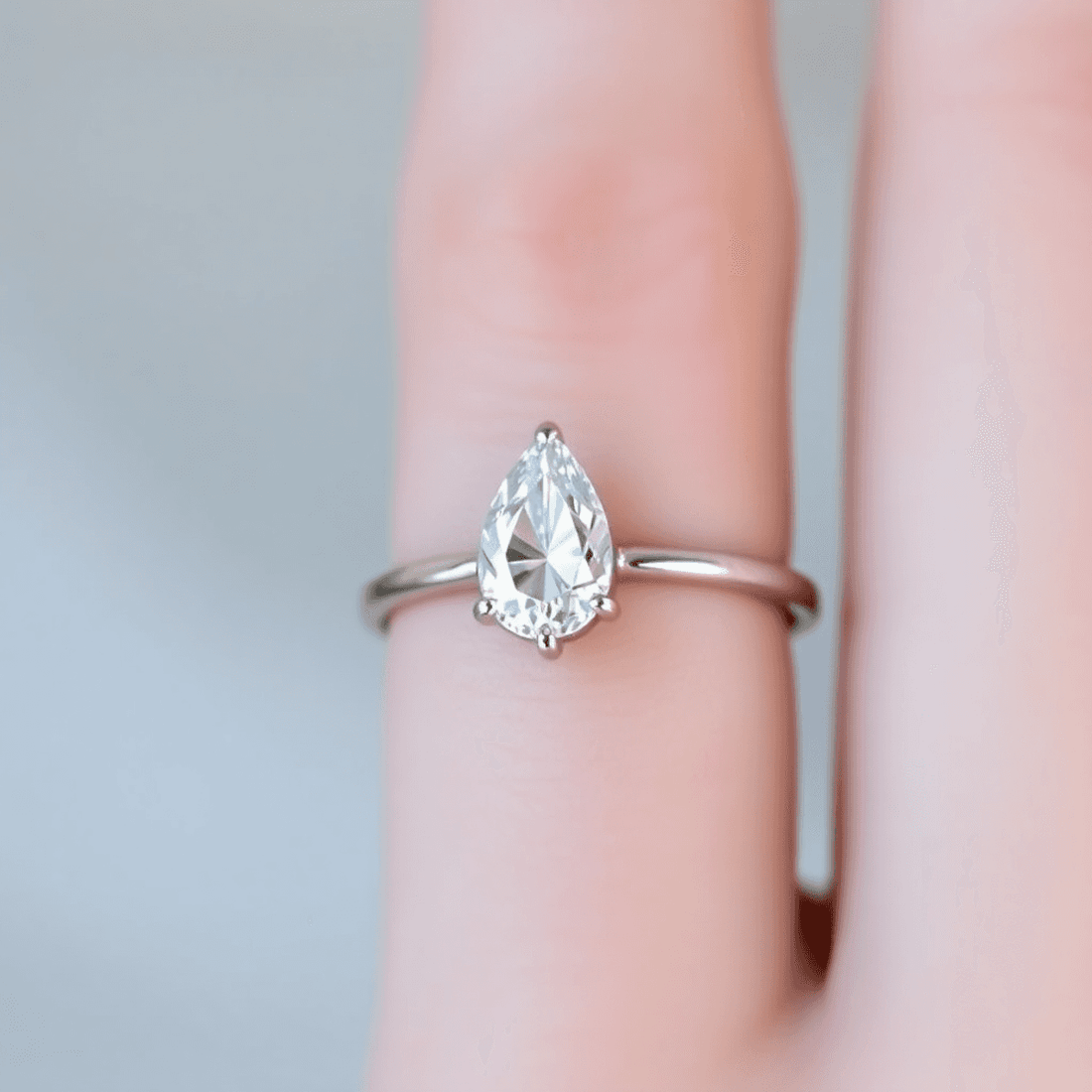 Close-up of a pear-shaped diamond engagement ring on a slender finger with a soft, blurred background highlighting its sparkle and elegant teardrop shape.