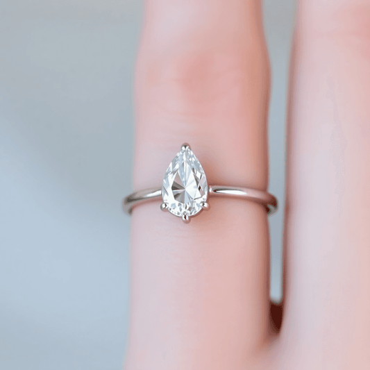 Close-up of a pear-shaped diamond engagement ring on a slender finger with a soft, blurred background highlighting its sparkle and elegant teardrop shape.