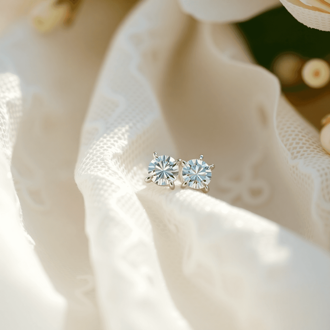 Close-up of sparkling diamond stud earrings on white lace fabric with soft flowers and gentle light, evoking bridal elegance and timeless sophistication.