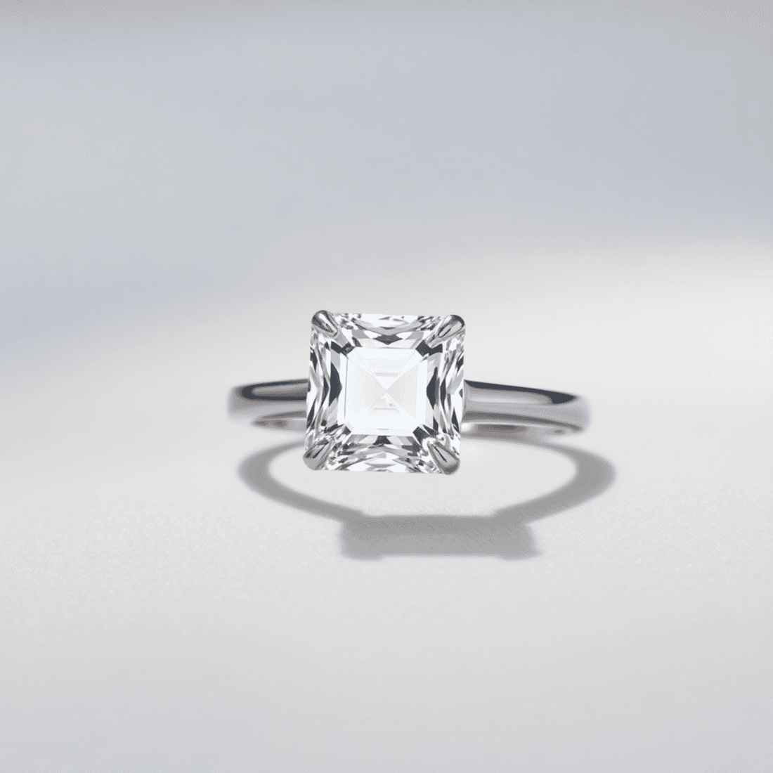 Close-up of a sparkling Asscher cut diamond ring on a sleek band, set against a soft neutral background with gentle light reflections highlighting its facets.