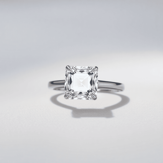 Close-up of a sparkling Asscher cut diamond ring on a sleek band, set against a soft neutral background with gentle light reflections highlighting its facets.
