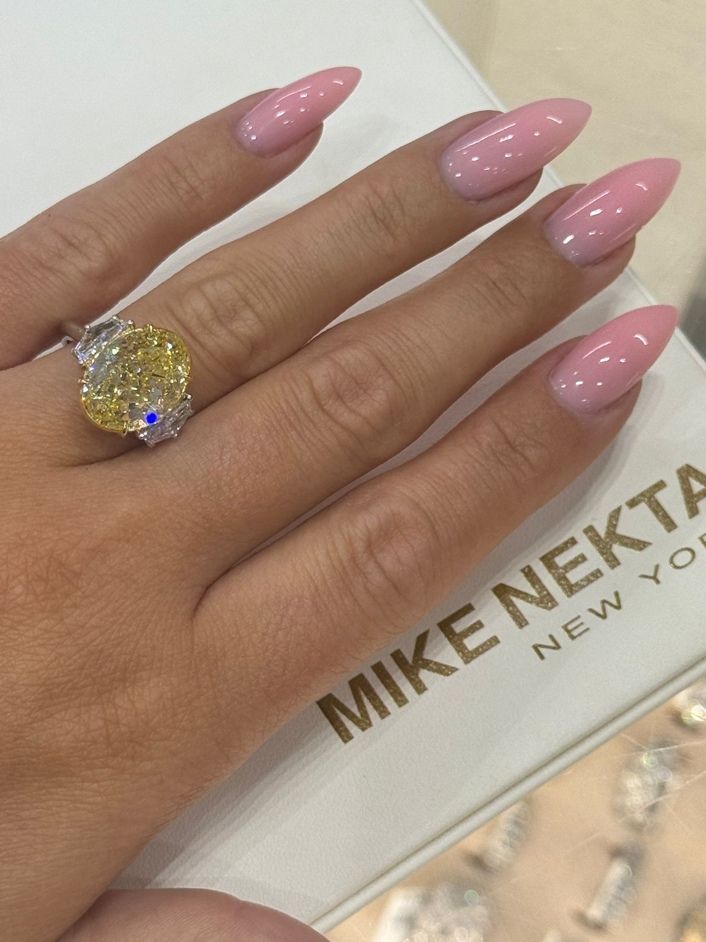 Hand with pink nails wearing a large diamond ring on a white background with 'MIKE NEKTA New York' branding.