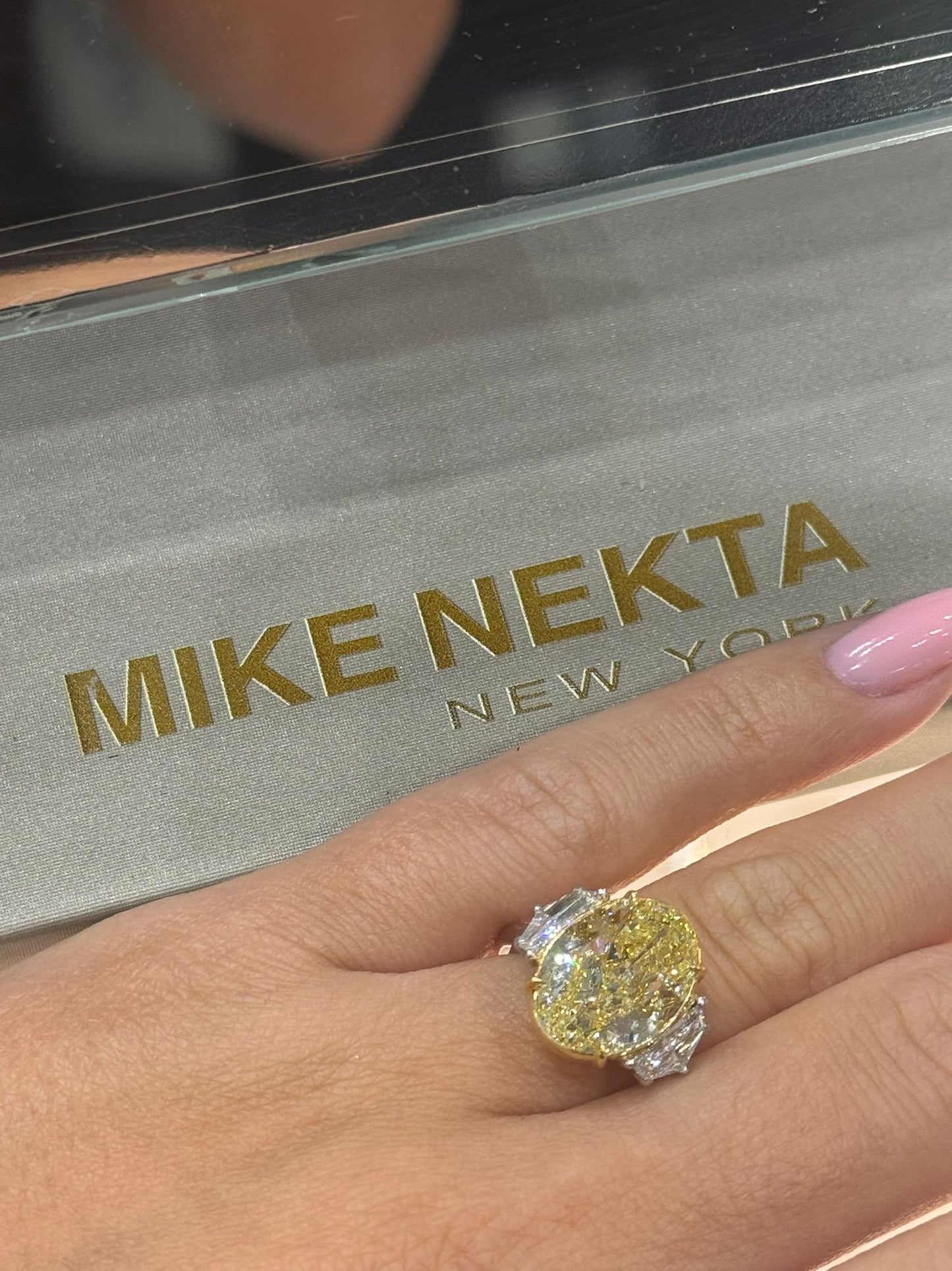 Gold ring with a large diamond on a hand, with 'MIKE NEKTA New York' branding in the background.