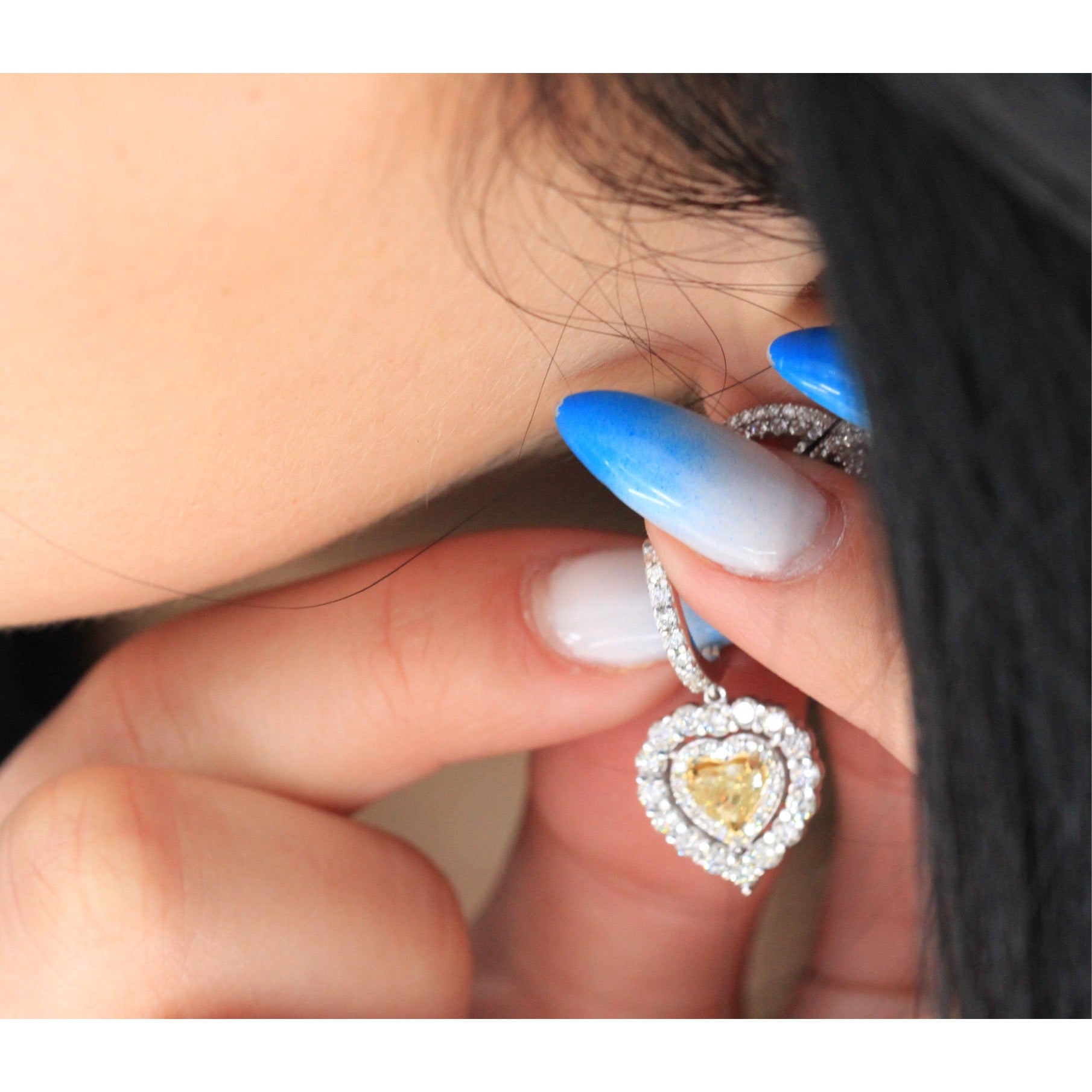 Yellow diamond heart in the double halo has an earring design being put on earlobe with manicured fingertips